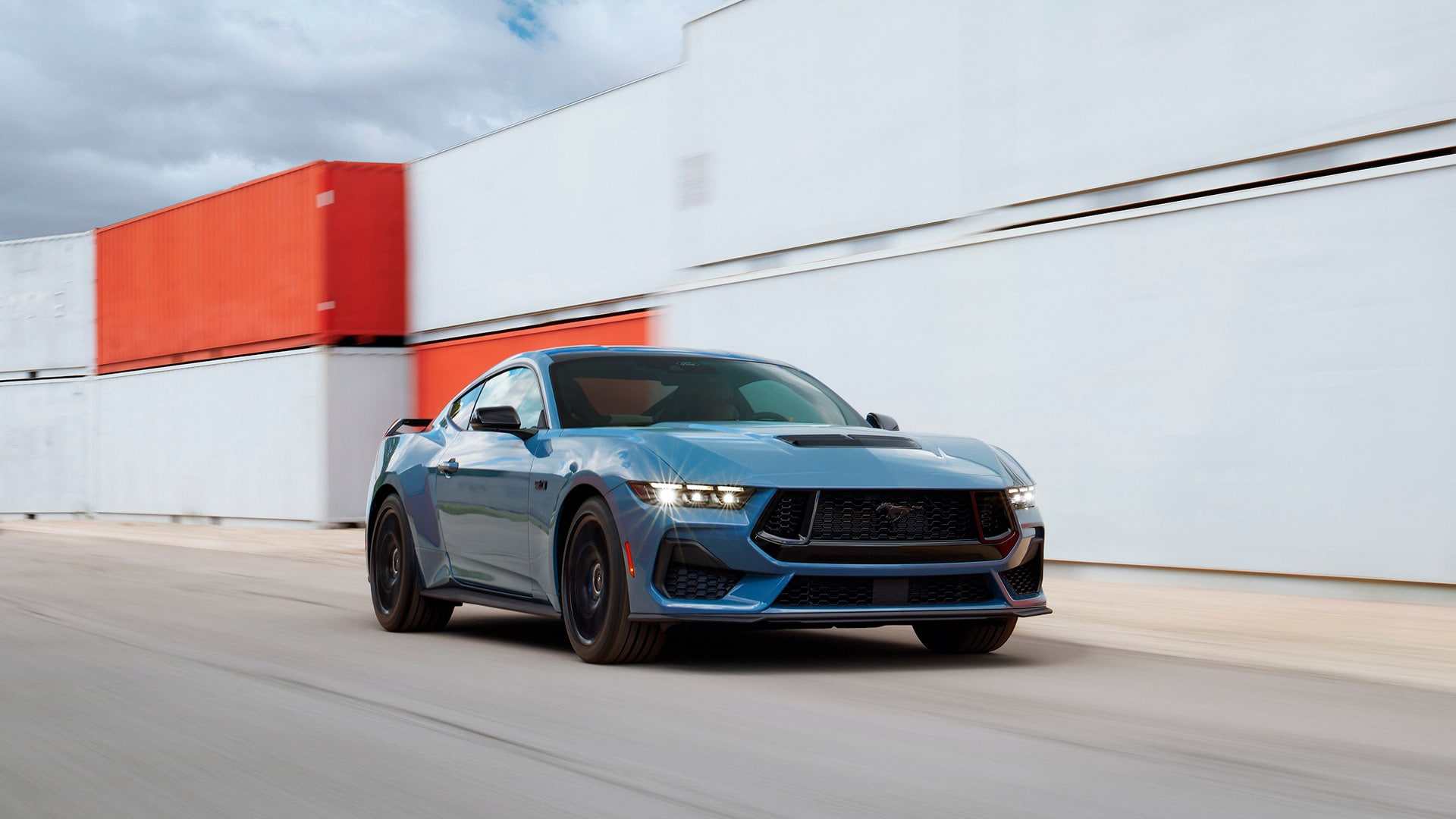 Ford Mustang GT azul em movimento, com contêineres ao fundo.