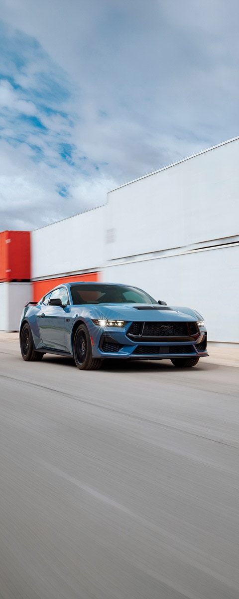 Ford Mustang GT azul em movimento, com contêineres ao fundo.